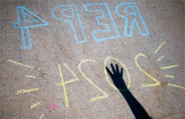 A person's hand showing the number 4 casts a shadow on sidewalk chalk that reads REP4 2024.