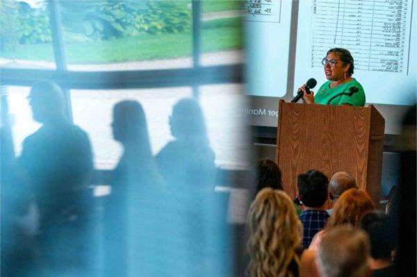  A person speaks at a lectern as people are reflected in glass nearby.