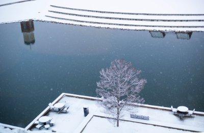 A snowy scene, seen from above. Looking down on snow covered tables, a tree, and a pond. The top of a building and carillon tower reflect on the pond.