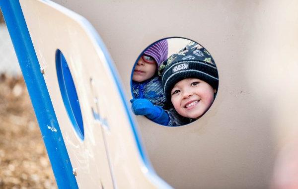 Two children look through a round opening of a piece of playground equipment at the camera. 