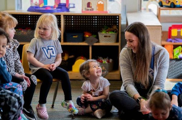 A preschool teacher and a young child smile at each other while sitting in a circle among other young children in a preschool classroom.