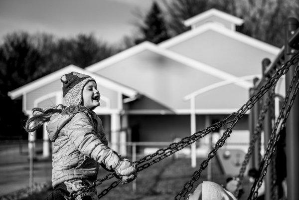 A little girl with a long ponytail and winter hat laughs as she swings on playground equipment outside a preschool.