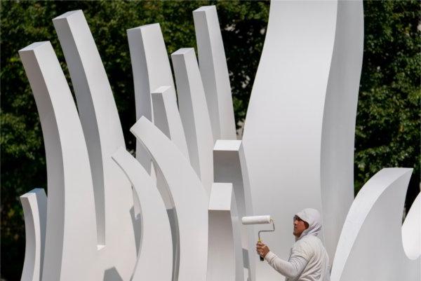 A person wearing white uses a paint roller to paint a metal art sculpture white. 