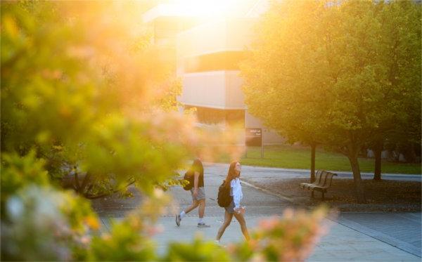  Two students walk through a college campus during sunrise with backpacks on their backs. 