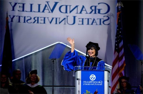 A university president wearing academic regalia waves to the audience while speaking at a lectern.  